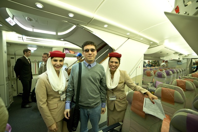 A young man stands between two girls dressed in Emirates corporate uniforms onboard A380, with stairs to the second floor of the aircraft visible in the background